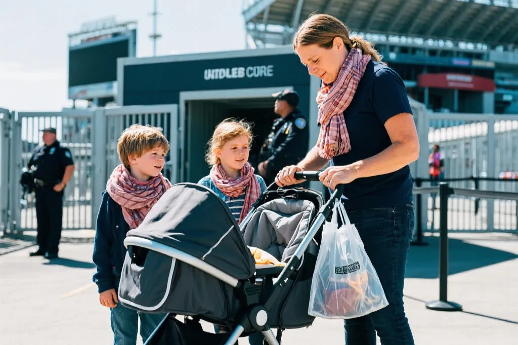 球场入口安检队伍中，家长推着可折叠婴儿车、背小包准备入场